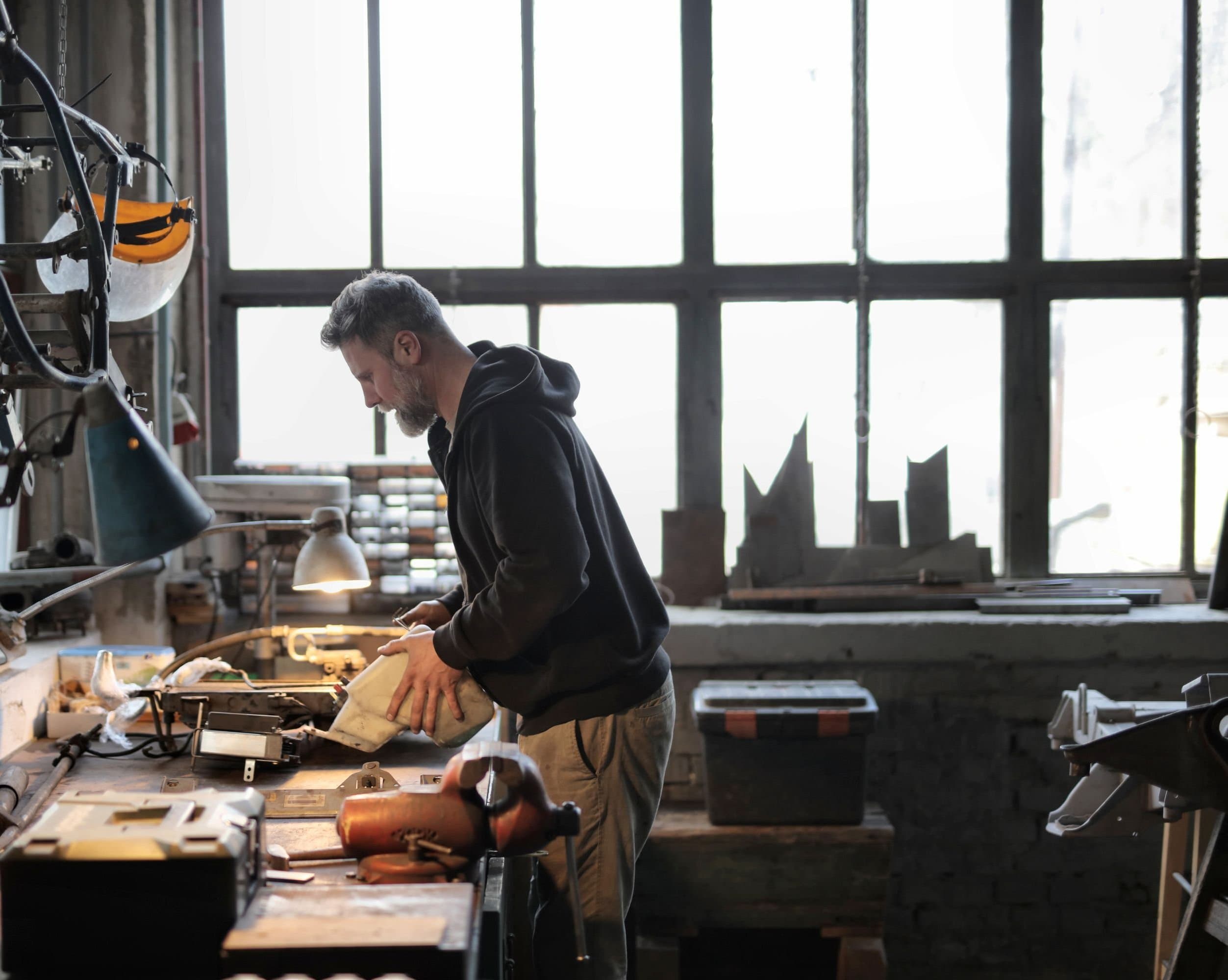 A man works at a bench under tall steel-framed windows in a machine shop.