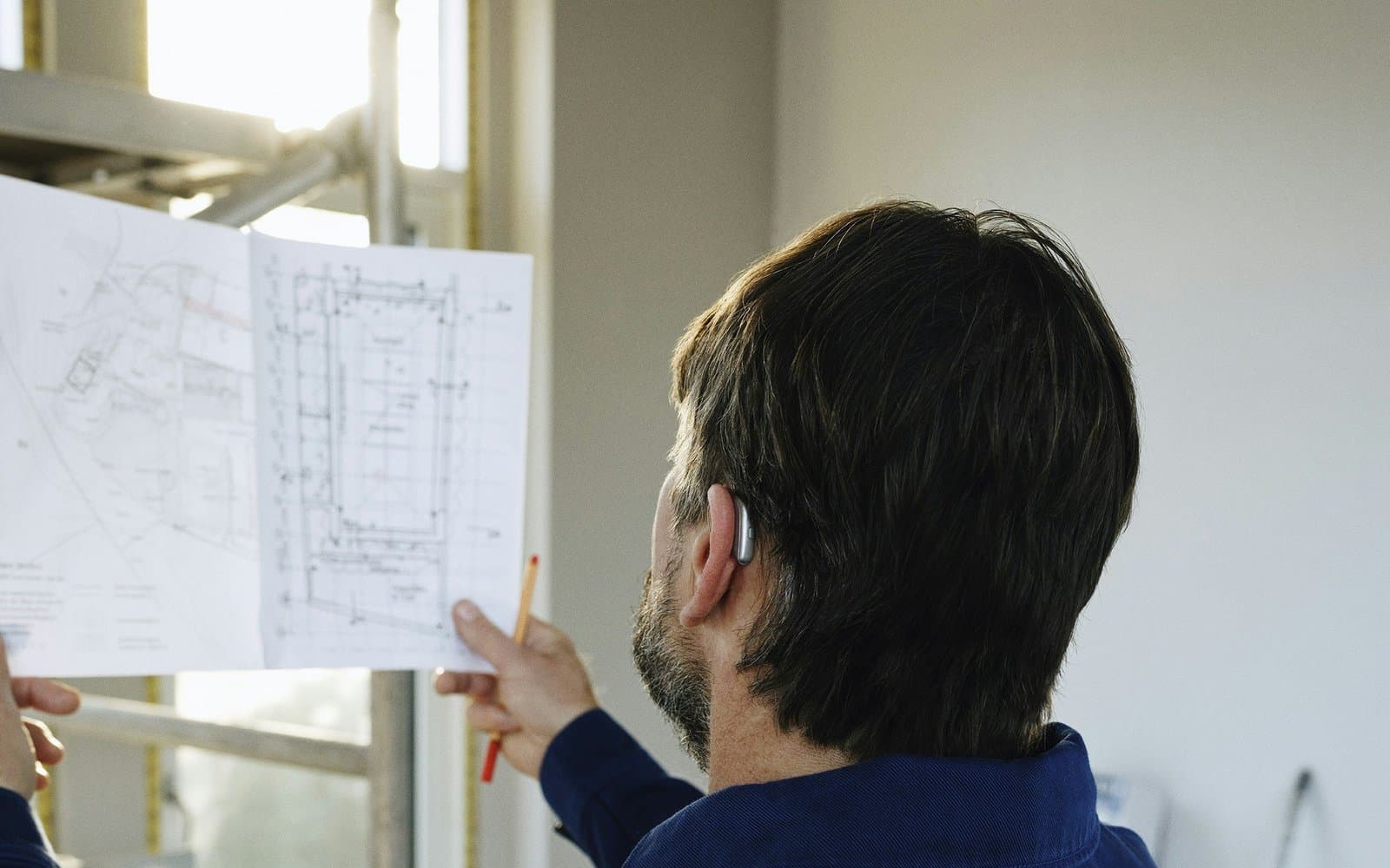 Seen from behind, a man in a navy work jacket studies a site plan in a building under renovation.