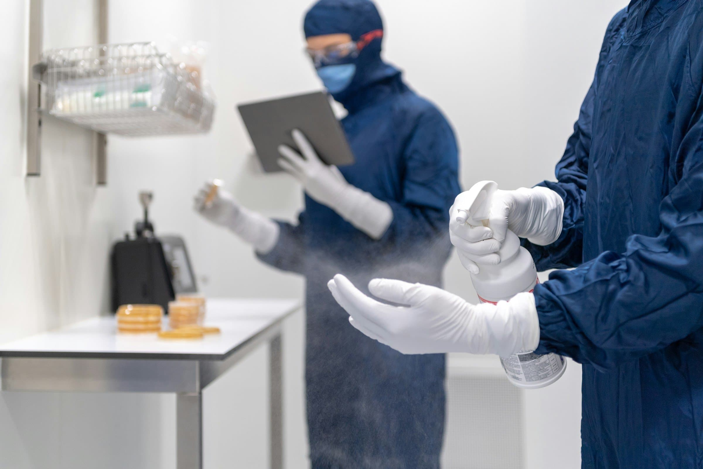 Two people in cleanroom gowns: one disinfects gloved hands while the other logs work on a tablet.